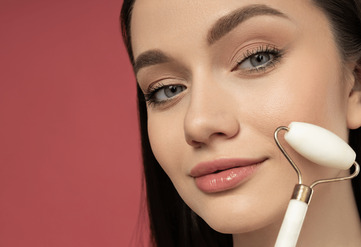 Close-up of a woman using a white jade roller on her cheek against a pink background.