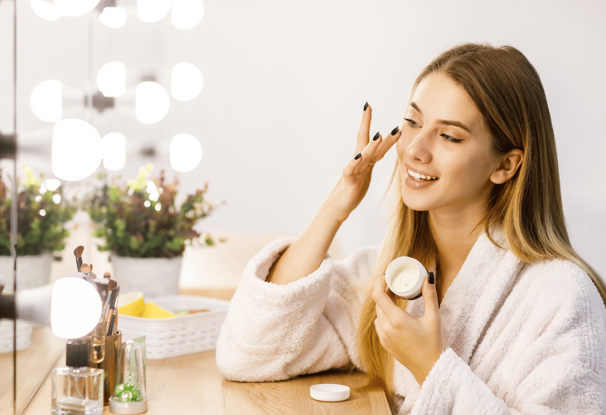 A woman sitting at her vanity, applying cream to her face in a cozy, warmly lit setting.