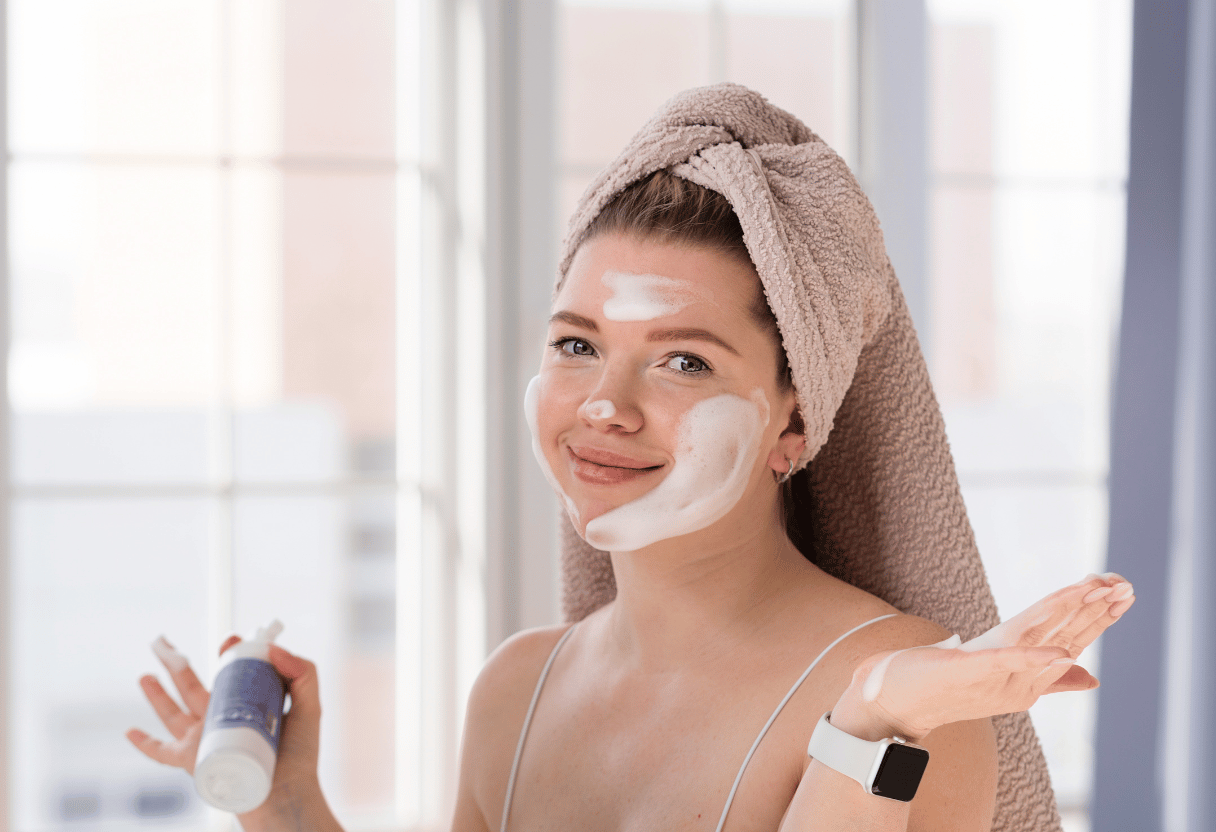 A young woman with a towel wrapped around her head, applying skincare cream to her face with a bright smile, in a softly lit room.