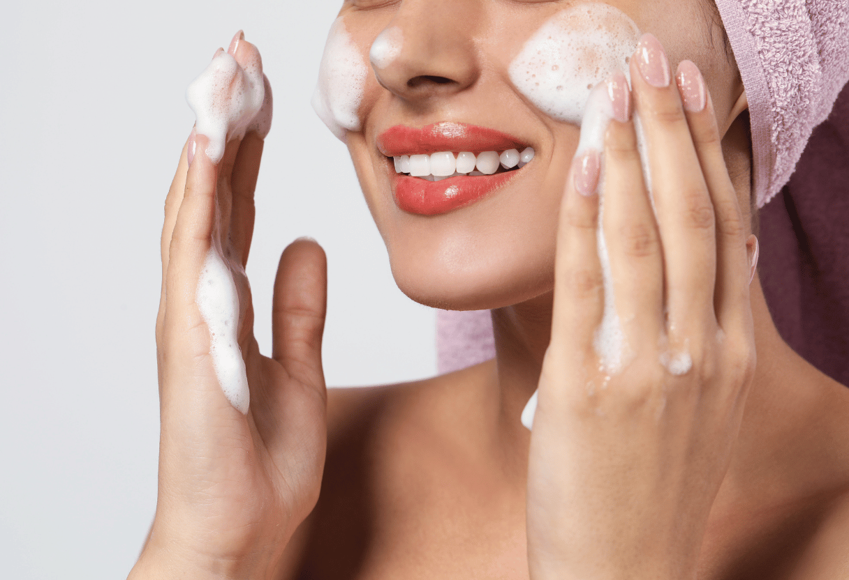 A close-up of a smiling woman with foam cleanser applied to her cheeks, emphasizing gentle cleansing.