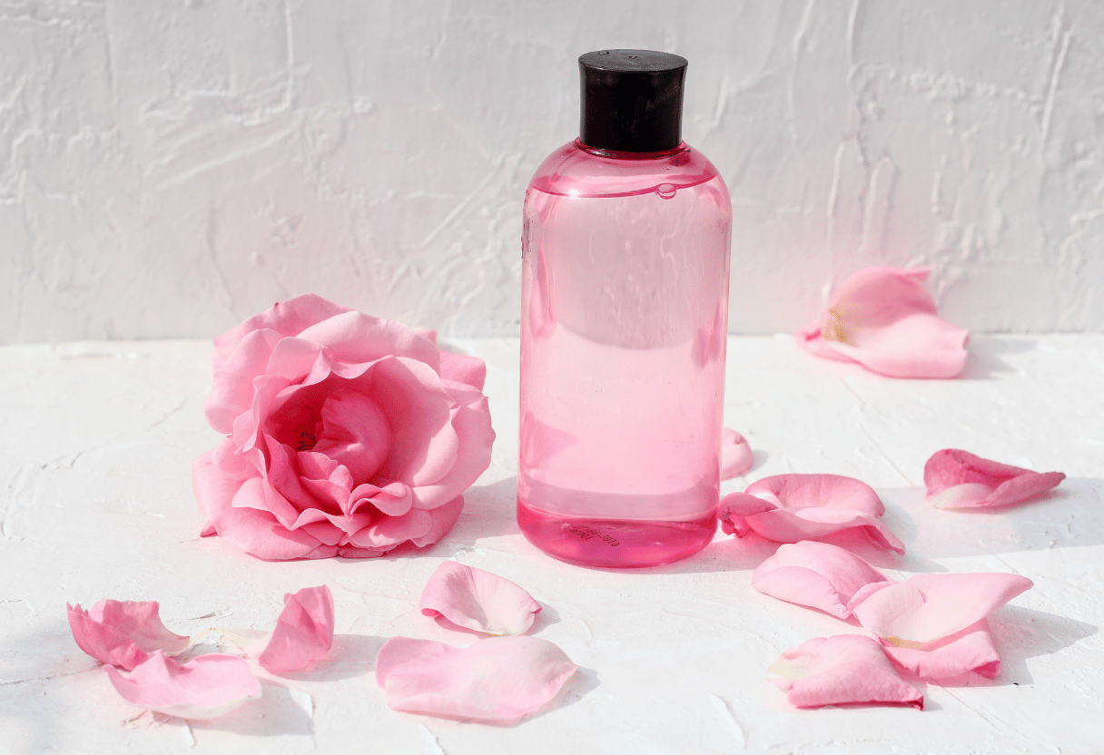 A pink rosewater bottle surrounded by rose petals on a white background