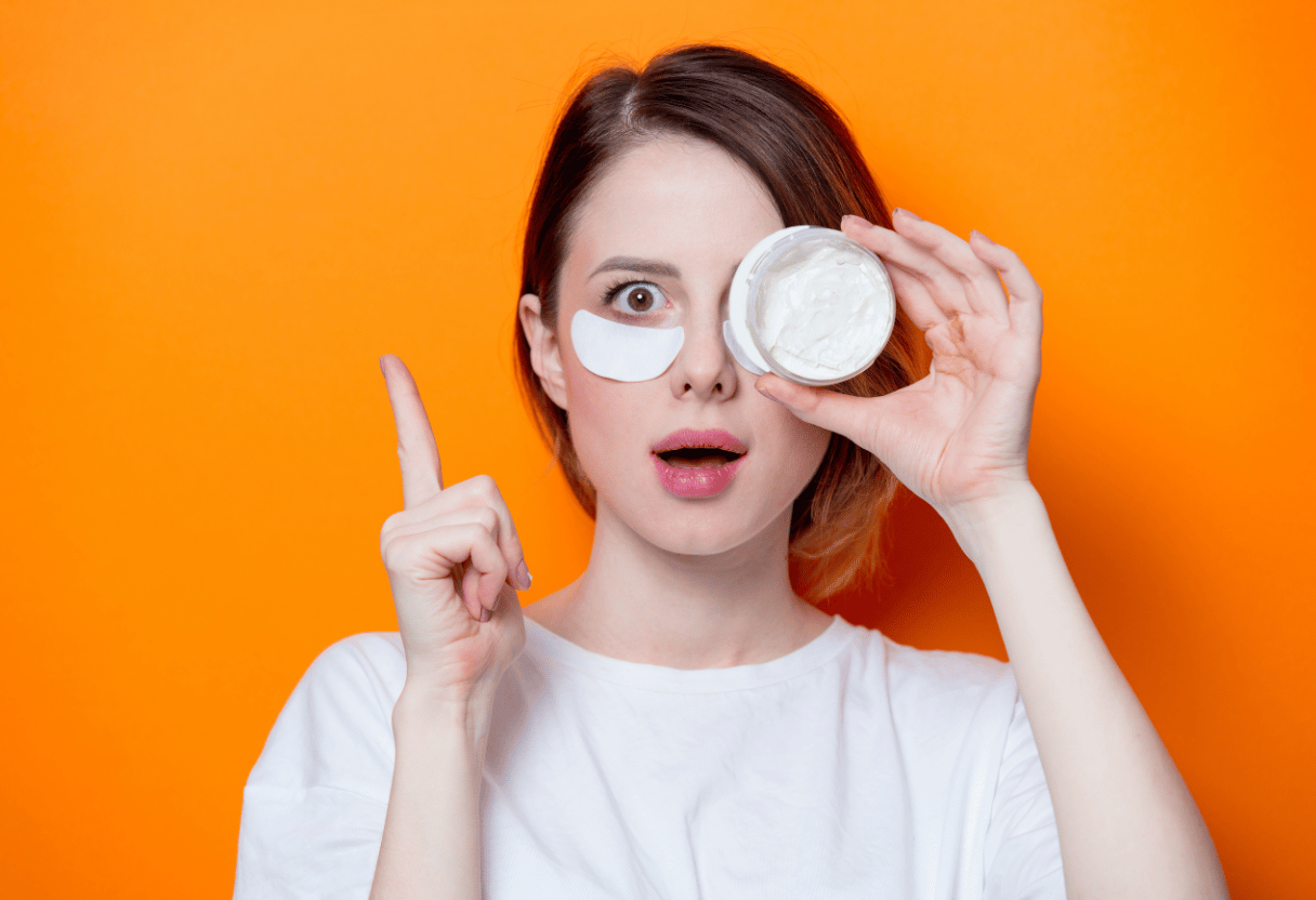 A woman holding a jar of cream and applying it near her eye with colorful orange background.