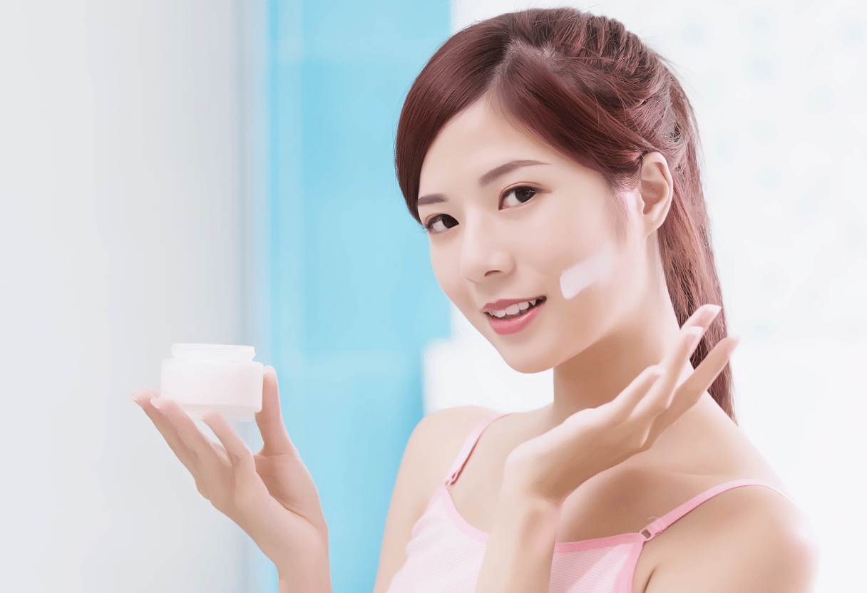 A woman smiling and applying moisturizer to her face with a background of a blue and white bathroom.