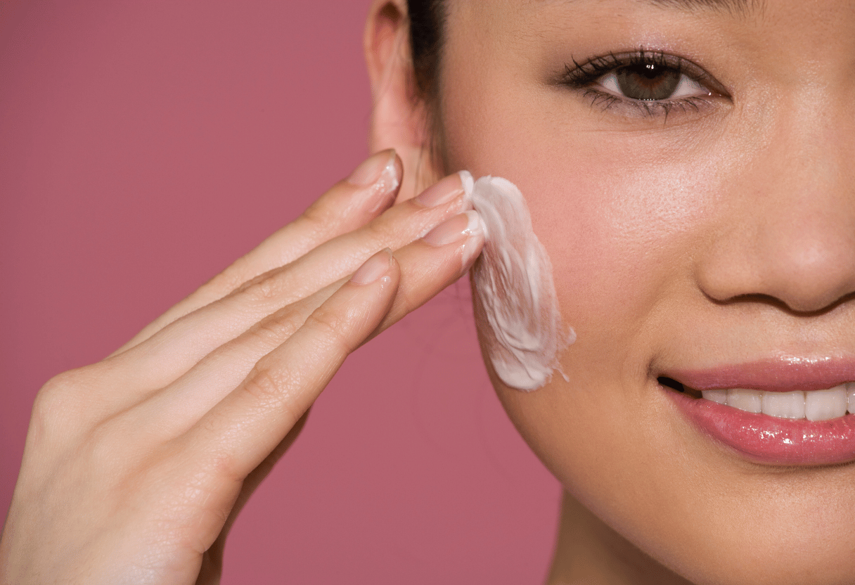 A close-up of a woman applying moisturizer to her cheek, set against a pink background.