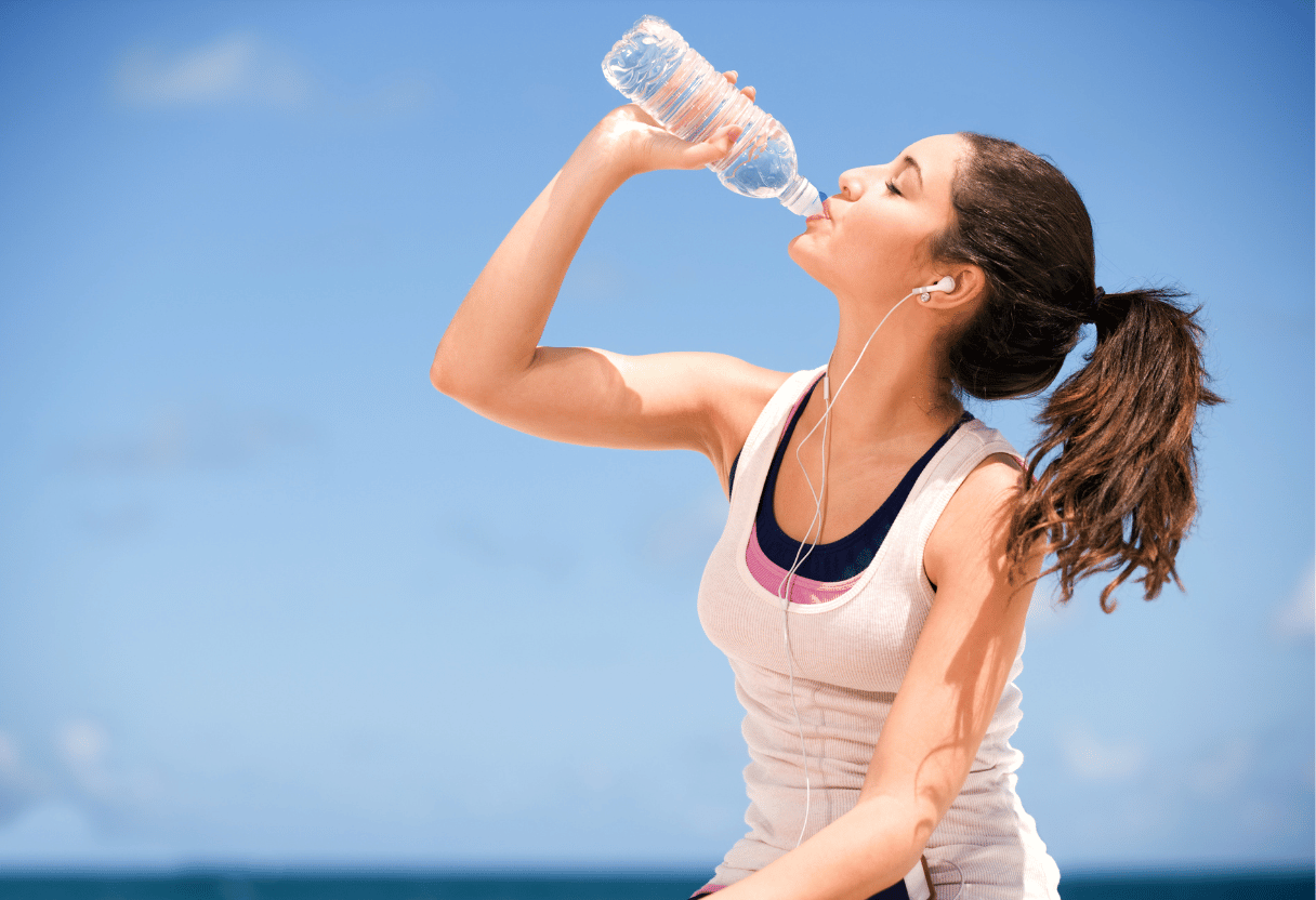 A fit woman drinking water from a bottle outdoors, wearing sportswear, under a clear blue sky.