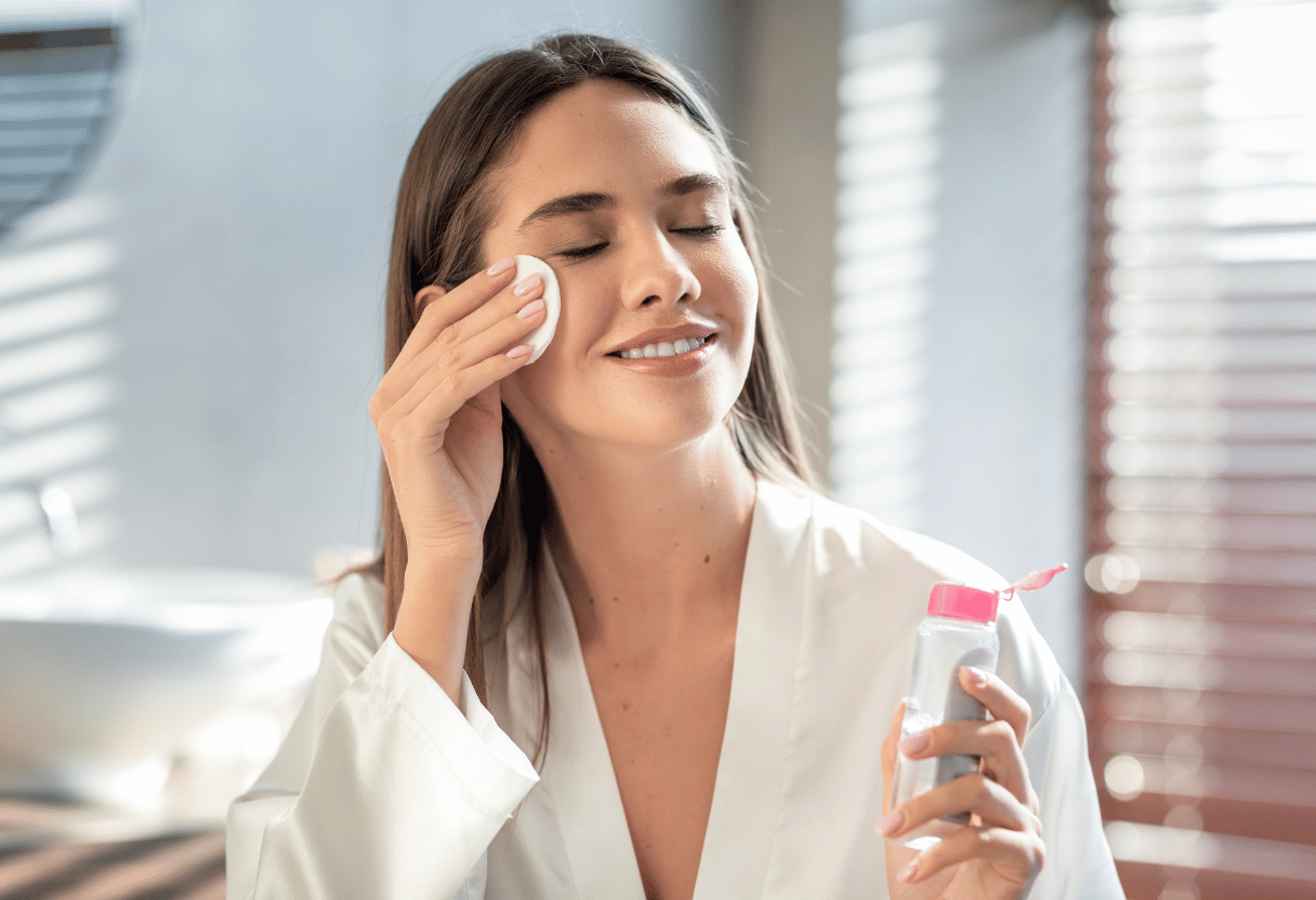 A woman in a white robe using a cotton pad to cleanse her face with a bottle of micellar water in hand.